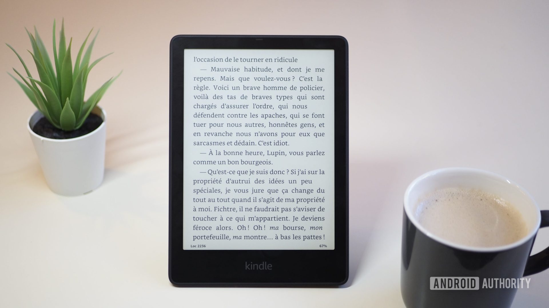 Kindle Paperwhite 2021 with the screen showing a book, on a table, next to a cup of coffee and plant, version 2