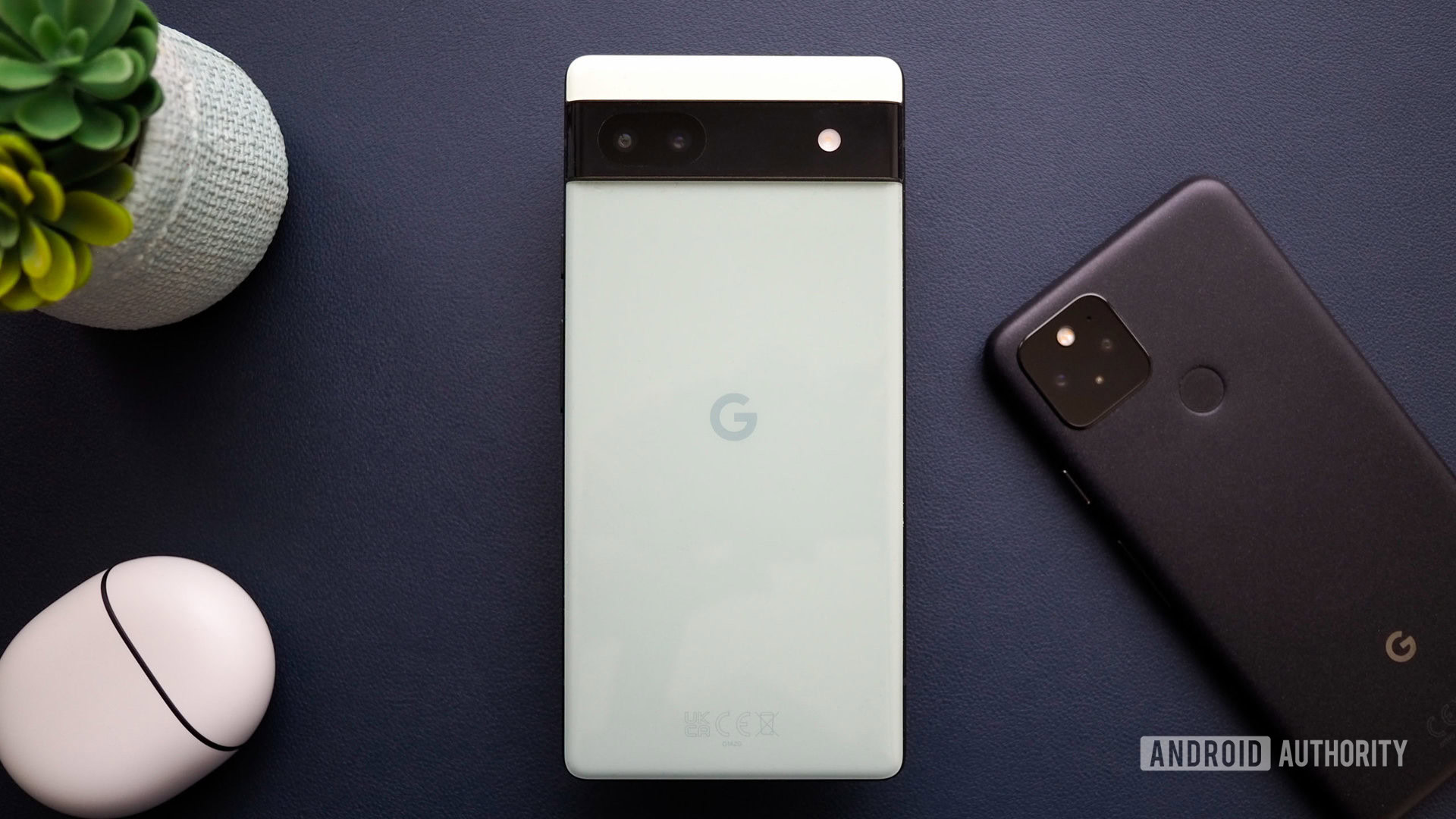 Google Pixel 6a in Sage color, seen from the back, next to a Pixel 5 and Pixel Buds Pro, on a navy background