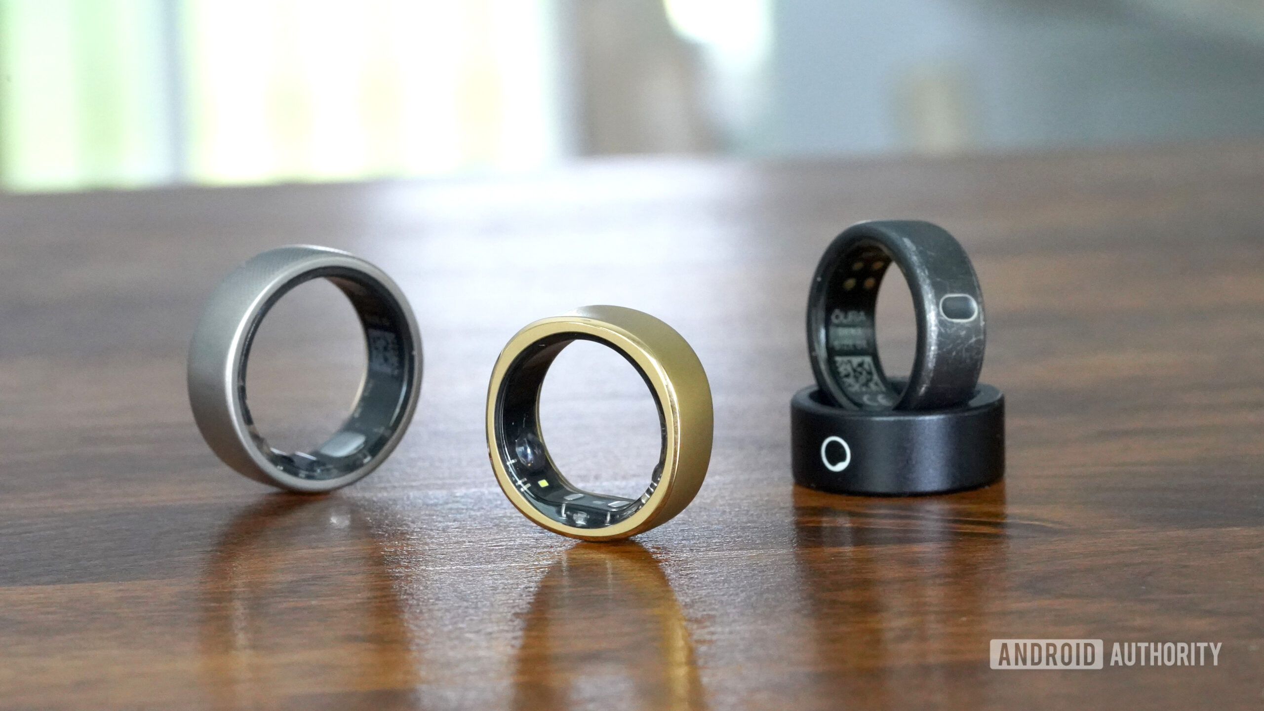 Une bague intelligente RingConn repose sur une surface en bois à côté d'une bague intelligente Oura Ring 3, Amazfit Hello et Circular.