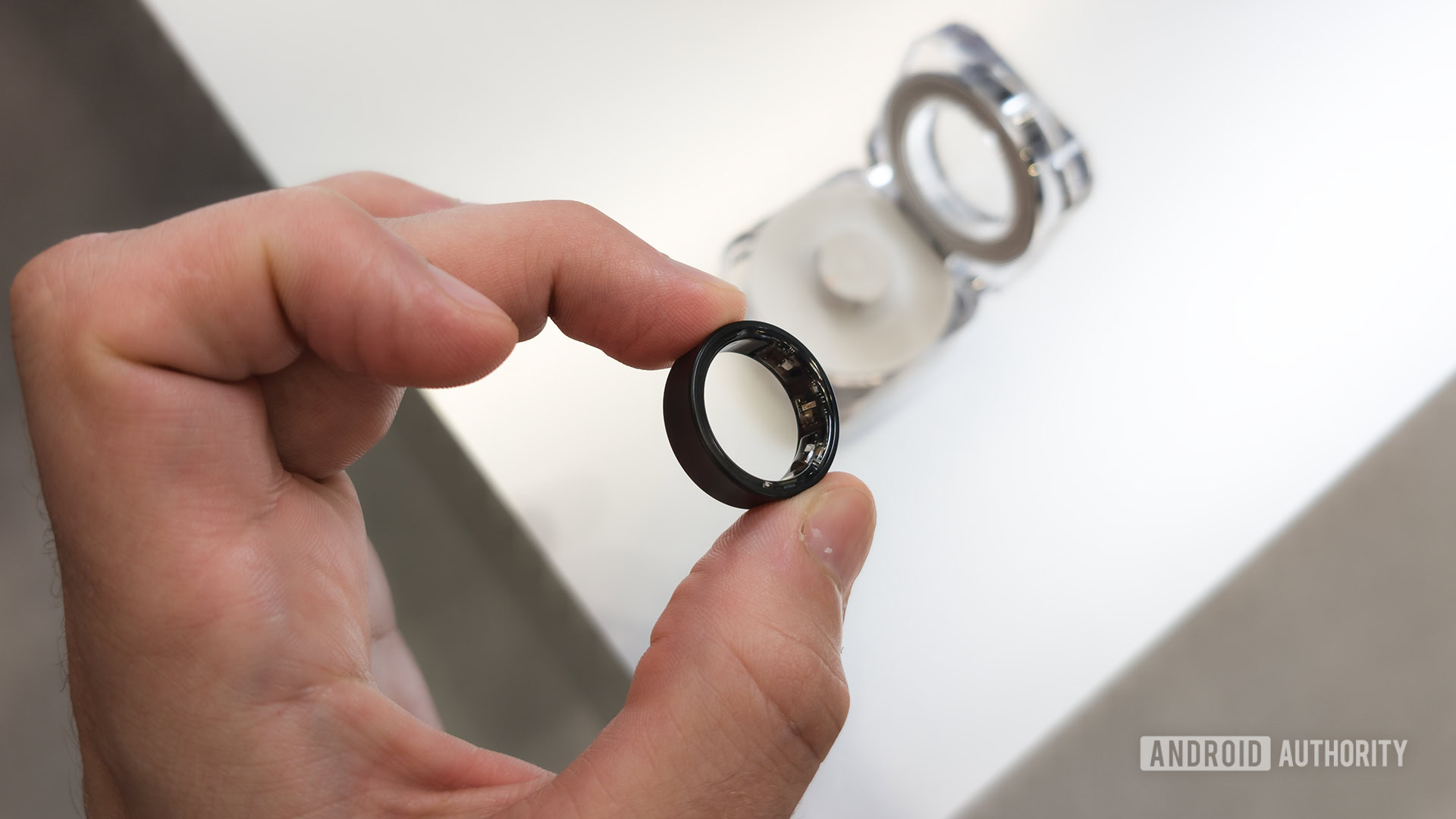 Samsung Galaxy Ring in Titanium Black Held Between Two Fingers with Case in Background
