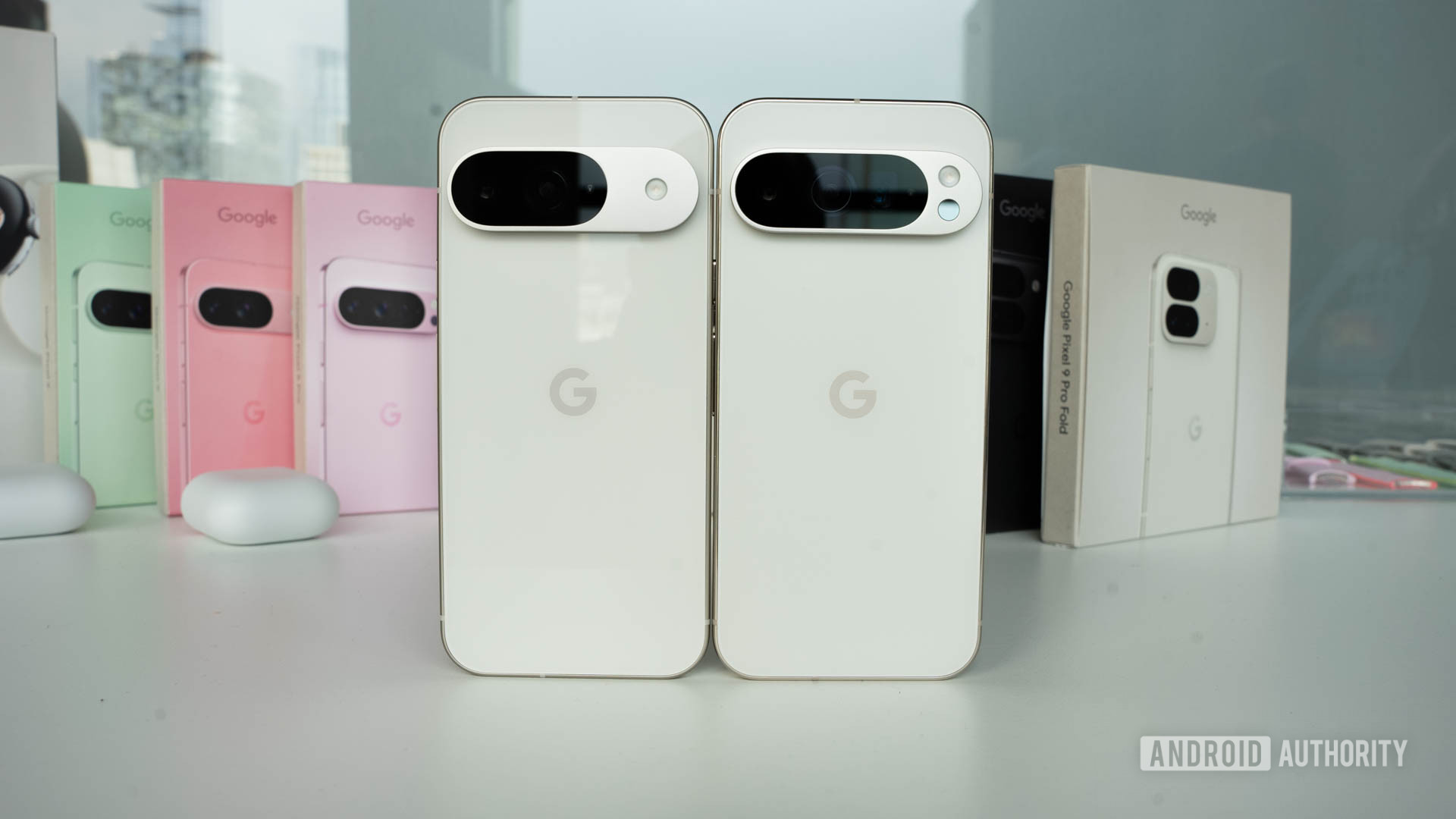 Google Pixel 9 in Porcelain standing next to Google Pixel 9 Pro in Porcelain with retail boxes in background