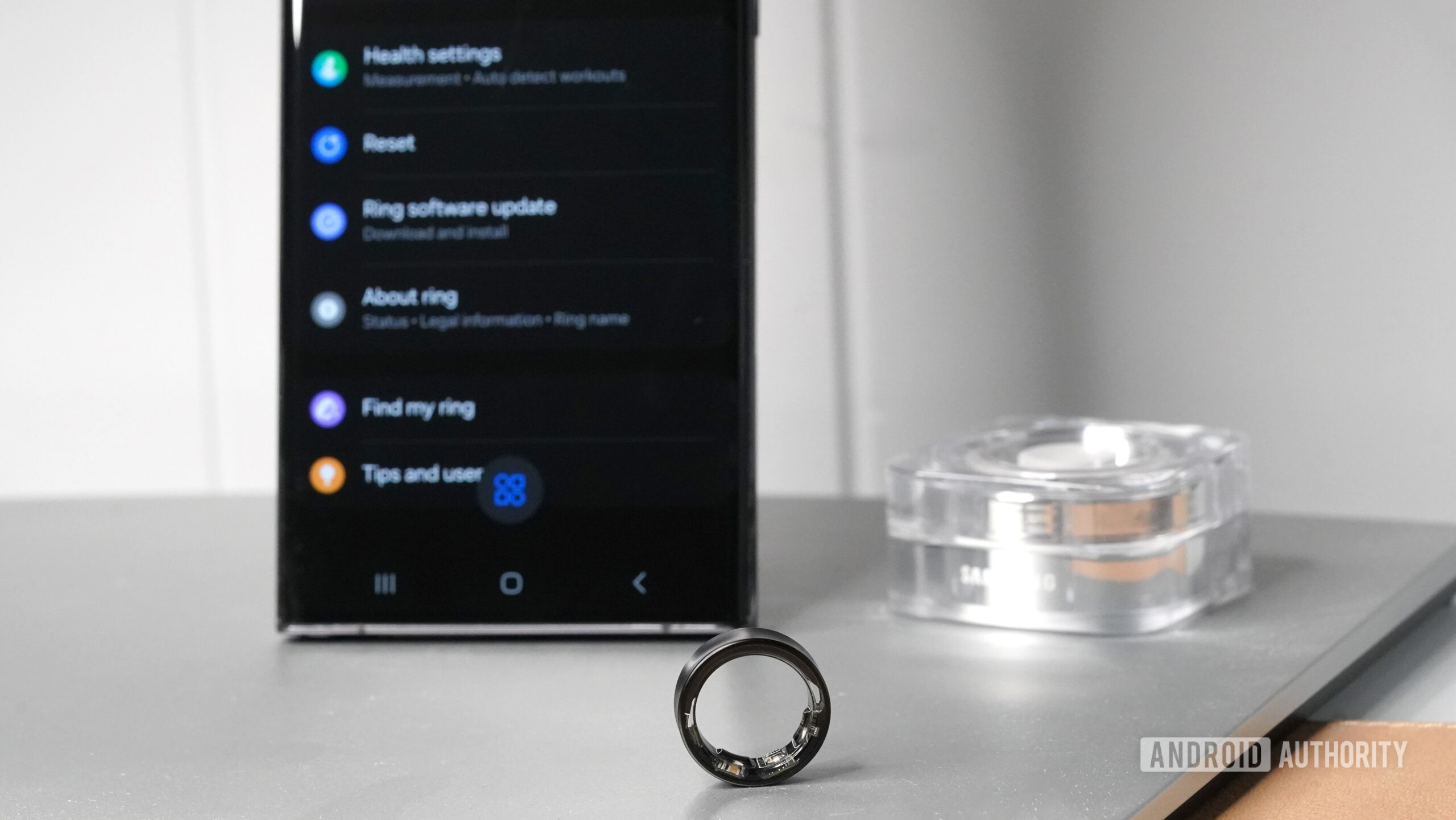 A Samsung Galaxy Ring rests in front of its charger, and a Samsung phone with the companion app displayed.