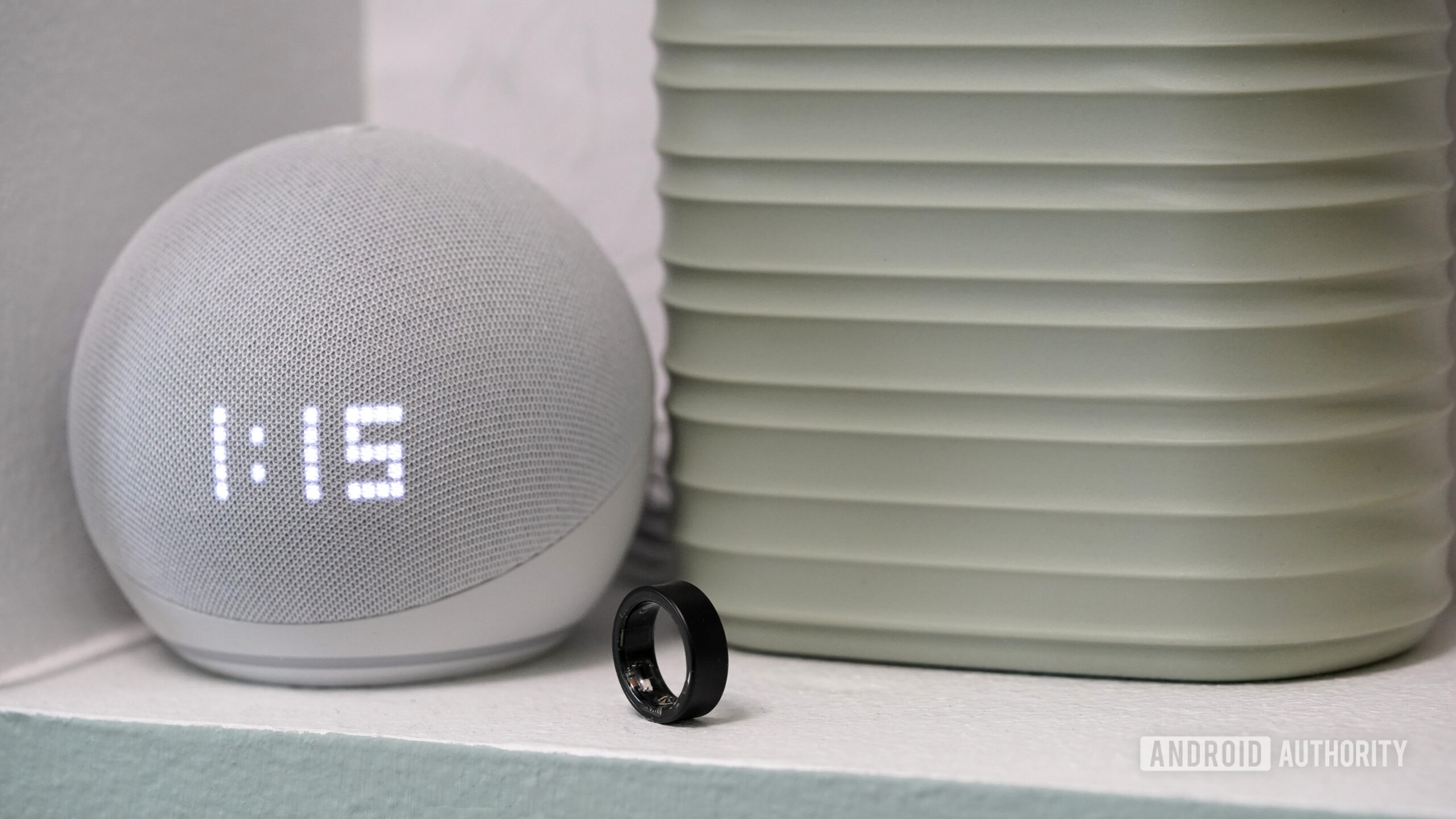 A Samsung Galaxy Ring rests next to a clock display 1:15 AM.