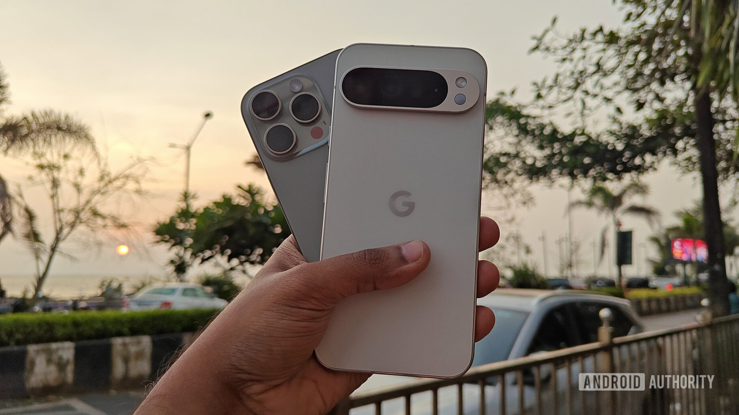 Apple iPhone 16 Pro and Google Pixel 9 Pro XL held out in hand, against the backdrop of a sunset on an urban promenade