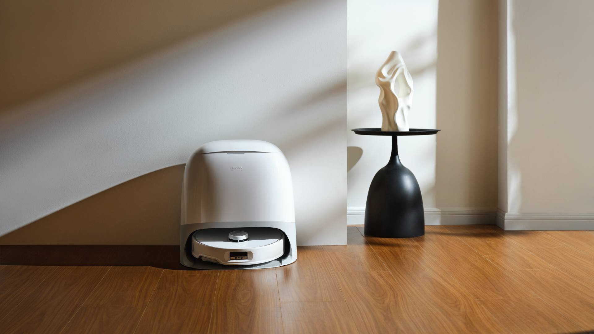 Roborock Qrevo Curv placed next to a wall on a wooden floor with a black coffe table to the side.